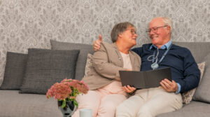 A couple sit on the sofa smiling at each other. The man is wearing a FlexO2 device and branded lanyard.
